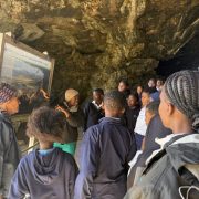 A group of school learners stands inside Klipgat Cave around a CapeNature interpretive board headed Life at Klipgat during the Middle Stone Age.