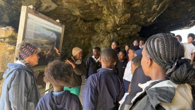 A group of school learners stands inside Klipgat Cave around a CapeNature interpretive board headed Life at Klipgat during the Middle Stone Age.