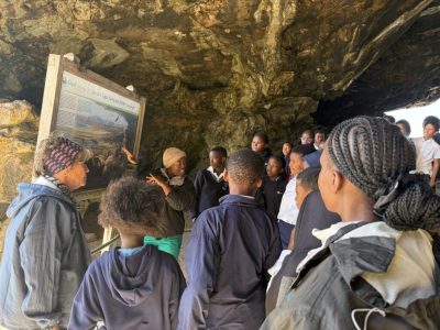 A group of school learners stands inside Klipgat Cave around a CapeNature interpretive board headed Life at Klipgat during the Middle Stone Age.