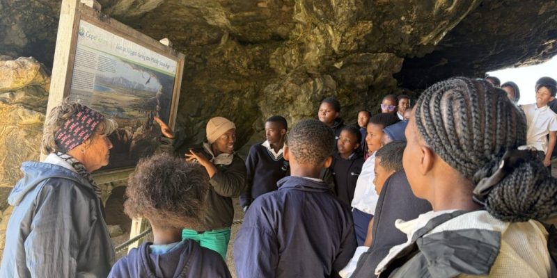 A group of school learners stands inside Klipgat Cave around a CapeNature interpretive board headed Life at Klipgat during the Middle Stone Age.