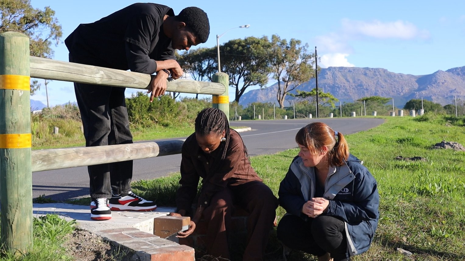 Three people at the roadside beside a toad tunnel entrance, one crouching with a camera-trap housing, a mountain ridge behind them.