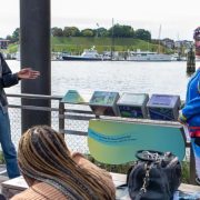 Two Baltimore Youth Arts apprentices speak to a seated audience on the National Aquarium Harbor Wetland boardwalk, beside a stand holding three illustrated tumbler panels, with the Inner Harbor behind them.