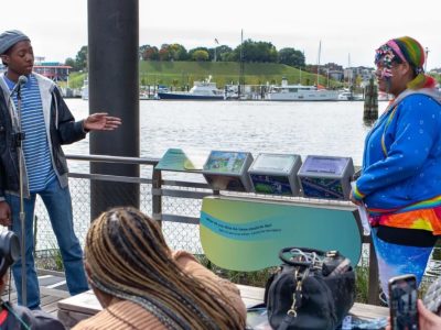 Two Baltimore Youth Arts apprentices speak to a seated audience on the National Aquarium Harbor Wetland boardwalk, beside a stand holding three illustrated tumbler panels, with the Inner Harbor behind them.