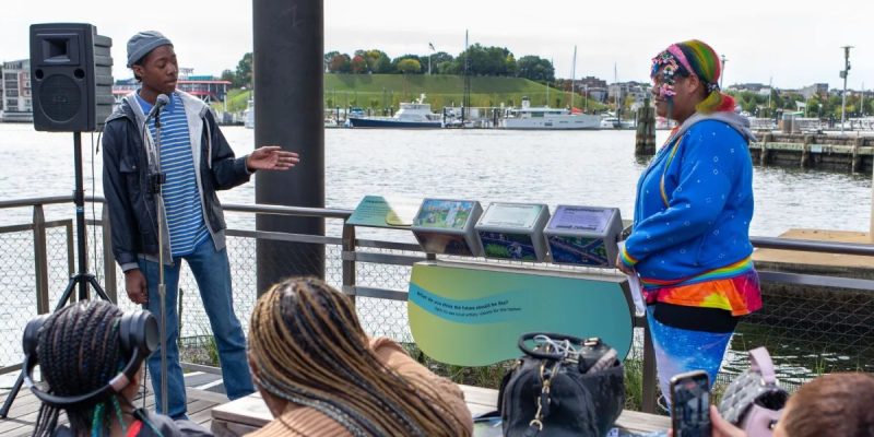 Two Baltimore Youth Arts apprentices speak to a seated audience on the National Aquarium Harbor Wetland boardwalk, beside a stand holding three illustrated tumbler panels, with the Inner Harbor behind them.