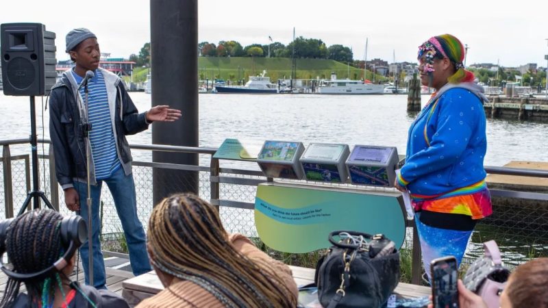 Two Baltimore Youth Arts apprentices speak to a seated audience on the National Aquarium Harbor Wetland boardwalk, beside a stand holding three illustrated tumbler panels, with the Inner Harbor behind them.