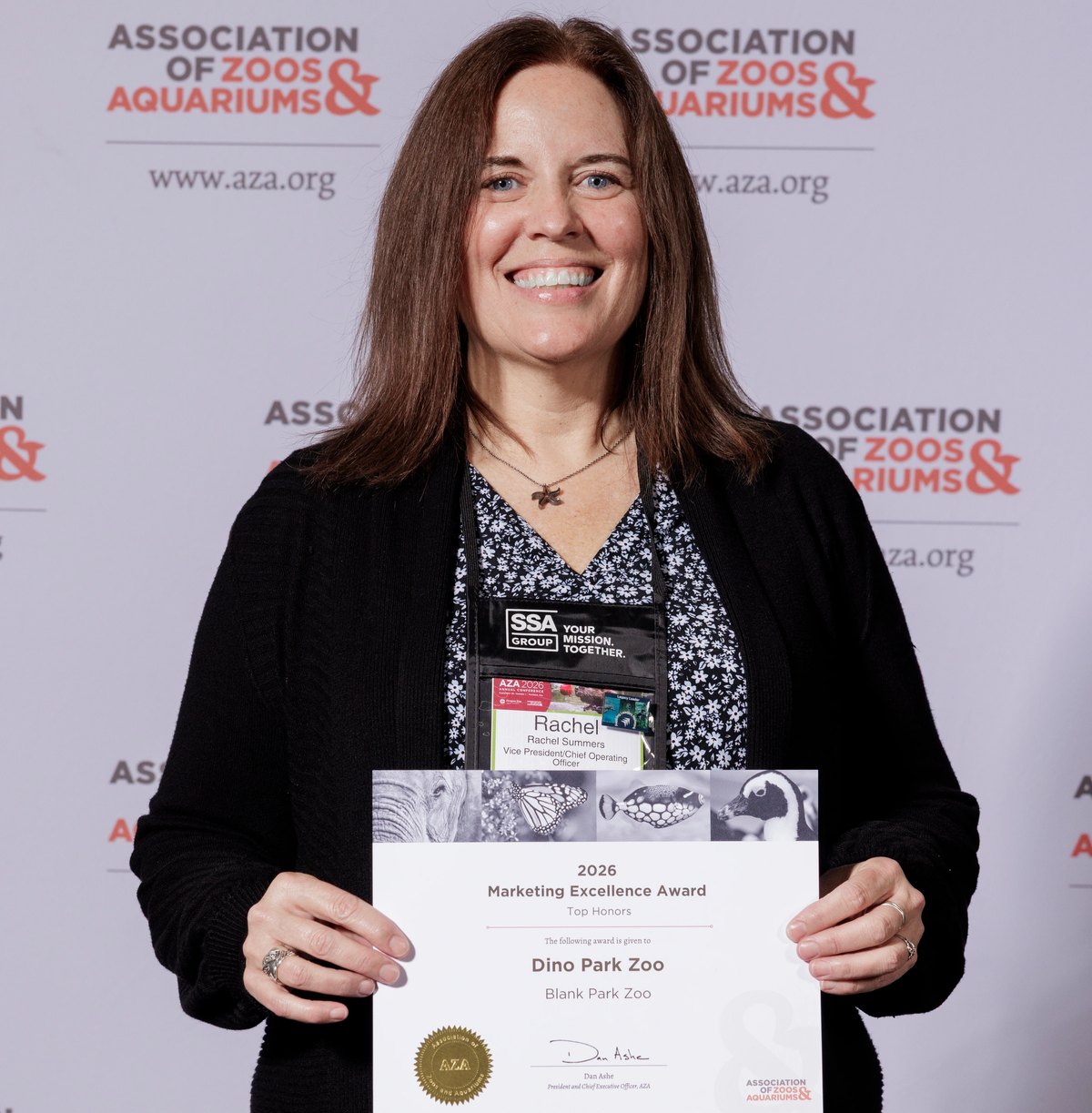 A Blank Park Zoo staff member in a black cardigan with a conference lanyard smiles while holding the zoo's award certificate up at chest height, standing in front of a backdrop printed repeatedly with the Association of Zoos and Aquariums logo.