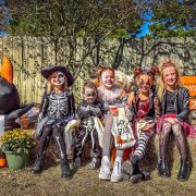 Five children in skeleton and cat Halloween costumes and face paint sit on hay bales at the Gulf Breeze Zoo, holding trick-or-treat bags, with an inflatable bat and pots of mums beside them.