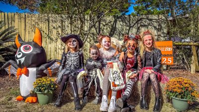 Five children in skeleton and cat Halloween costumes and face paint sit on hay bales at the Gulf Breeze Zoo, holding trick-or-treat bags, with an inflatable bat and pots of mums beside them.