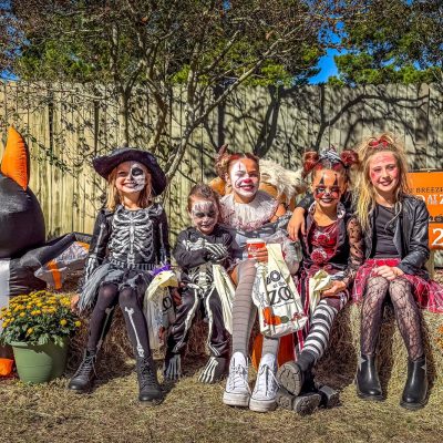 Five children in skeleton and cat Halloween costumes and face paint sit on hay bales at the Gulf Breeze Zoo, holding trick-or-treat bags, with an inflatable bat and pots of mums beside them.