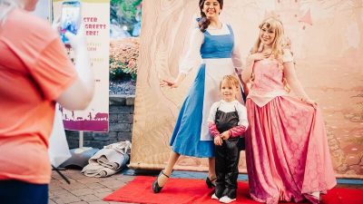 A young child in a black costume with white gloves stands on a red carpet between two costumed characters in long gowns, one blue and white and one pink, in front of a painted storybook castle backdrop, while an adult in an orange shirt photographs them with a phone beside a Boo at the Zoo character meet and greet sign.
