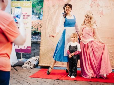 A young child in a black costume with white gloves stands on a red carpet between two costumed characters in long gowns, one blue and white and one pink, in front of a painted storybook castle backdrop, while an adult in an orange shirt photographs them with a phone beside a Boo at the Zoo character meet and greet sign.