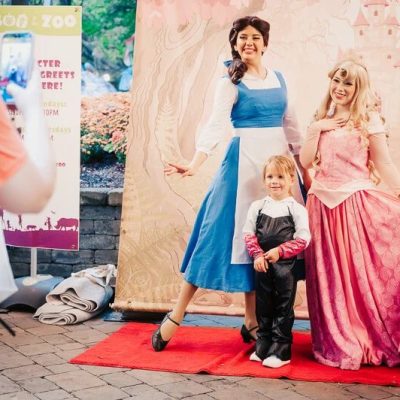 A young child in a black costume with white gloves stands on a red carpet between two costumed characters in long gowns, one blue and white and one pink, in front of a painted storybook castle backdrop, while an adult in an orange shirt photographs them with a phone beside a Boo at the Zoo character meet and greet sign.