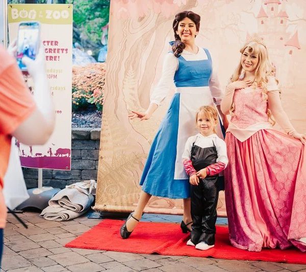 A young child in a black costume with white gloves stands on a red carpet between two costumed characters in long gowns, one blue and white and one pink, in front of a painted storybook castle backdrop, while an adult in an orange shirt photographs them with a phone beside a Boo at the Zoo character meet and greet sign.