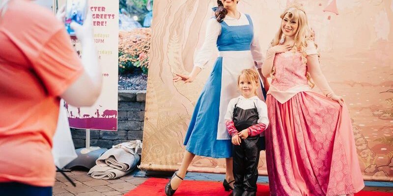 A young child in a black costume with white gloves stands on a red carpet between two costumed characters in long gowns, one blue and white and one pink, in front of a painted storybook castle backdrop, while an adult in an orange shirt photographs them with a phone beside a Boo at the Zoo character meet and greet sign.