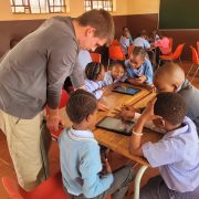 A facilitator leans over a classroom desk where primary school learners in uniform work together around two digital tablets, part of a conservation education program in Mpumalanga, South Africa.