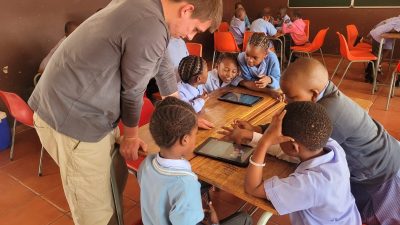 A facilitator leans over a classroom desk where primary school learners in uniform work together around two digital tablets, part of a conservation education program in Mpumalanga, South Africa.