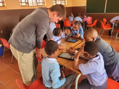A facilitator leans over a classroom desk where primary school learners in uniform work together around two digital tablets, part of a conservation education program in Mpumalanga, South Africa.