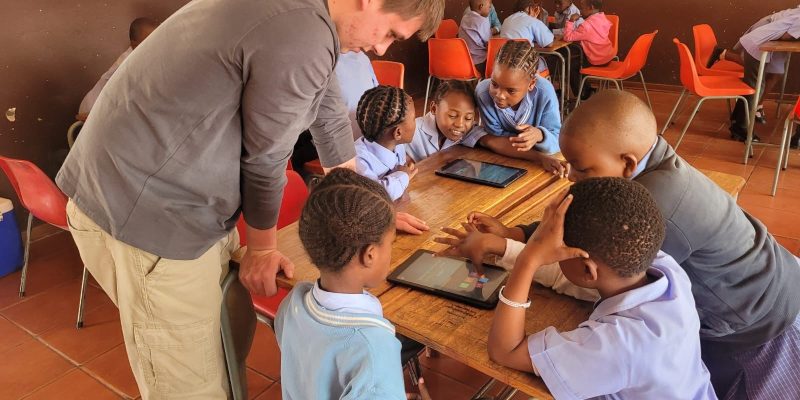 A facilitator leans over a classroom desk where primary school learners in uniform work together around two digital tablets, part of a conservation education program in Mpumalanga, South Africa.