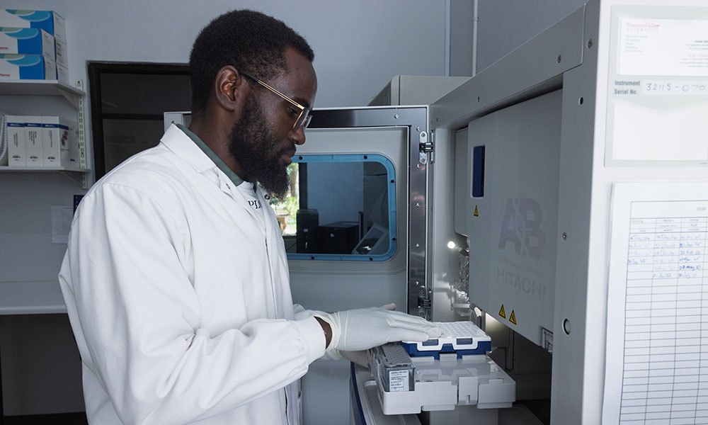 A scientist in a white laboratory coat and gloves loads a tray of sample tubes into an open benchtop genetic analyzer, with reagent shelves and a printed log sheet on the wall beside him.