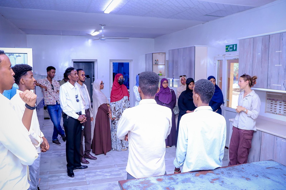 Students from the University of Hargeisa stand in a tiled treatment room at the Cheetah Conservation Fund's Somaliland sanctuary, listening to a CCF staff member in a khaki field shirt who is standing beside a counter below a wall chart.