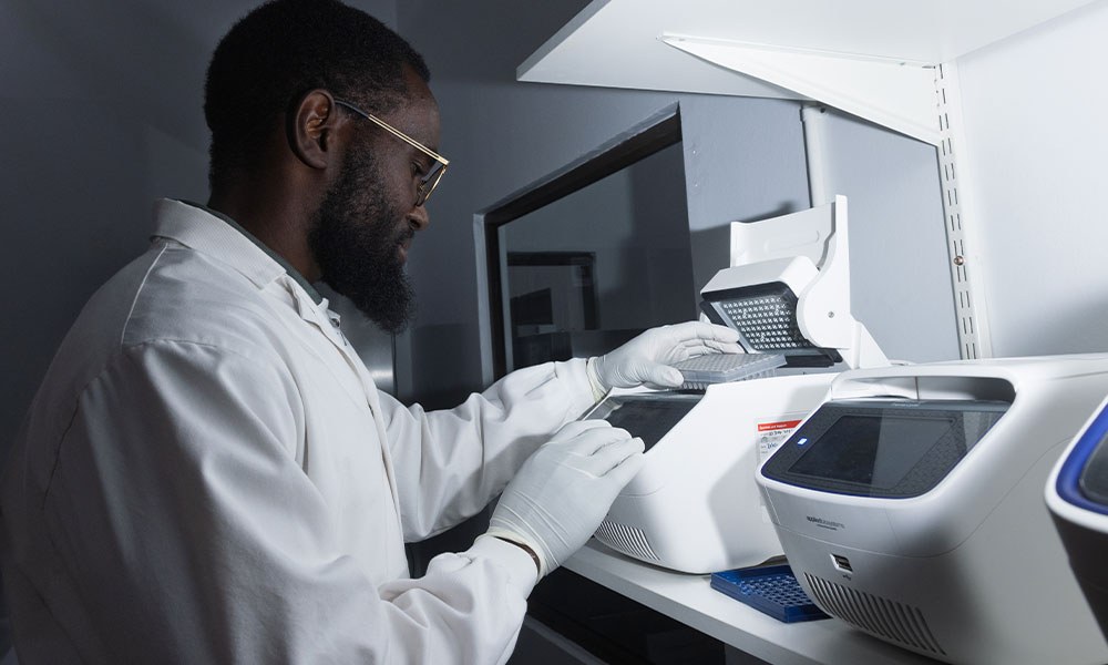 A scientist in a white coat and gloves loads a sample plate into a thermal cycler at the Cheetah Conservation Fund genetics laboratory in Namibia
