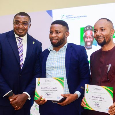 Three men stand for a photograph at the Chief S.L. Edu Memorial Lecture, two of them holding Nigerian Conservation Foundation research grant certificates, in front of a backdrop carrying the foundation's logo and the lecture title.