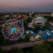 An aerial view at dusk of the Downtown Aquarium Houston complex, with a large Ferris wheel lit in bands of pink, green and blue on the left, the aquarium building marked with a large AQUARIUM sign at right, blue-roofed pavilions and palms between them, and a freeway curving past behind.