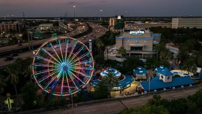 An aerial view at dusk of the Downtown Aquarium Houston complex, with a large Ferris wheel lit in bands of pink, green and blue on the left, the aquarium building marked with a large AQUARIUM sign at right, blue-roofed pavilions and palms between them, and a freeway curving past behind.