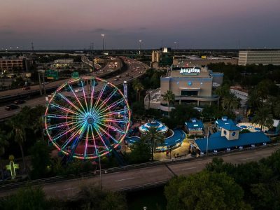 An aerial view at dusk of the Downtown Aquarium Houston complex, with a large Ferris wheel lit in bands of pink, green and blue on the left, the aquarium building marked with a large AQUARIUM sign at right, blue-roofed pavilions and palms between them, and a freeway curving past behind.