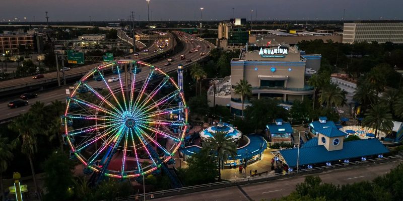 An aerial view at dusk of the Downtown Aquarium Houston complex, with a large Ferris wheel lit in bands of pink, green and blue on the left, the aquarium building marked with a large AQUARIUM sign at right, blue-roofed pavilions and palms between them, and a freeway curving past behind.