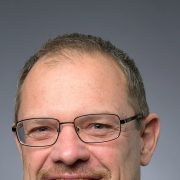 Dwight Scott, president and CEO of the Saint Louis Zoo, in a studio portrait wearing a dark suit, white shirt and blue patterned tie, with glasses and a short gray beard, against a gray background.