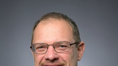 Dwight Scott, president and CEO of the Saint Louis Zoo, in a studio portrait wearing a dark suit, white shirt and blue patterned tie, with glasses and a short gray beard, against a gray background.