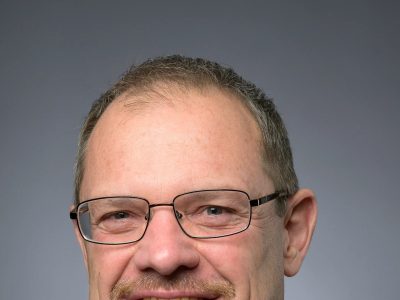Dwight Scott, president and CEO of the Saint Louis Zoo, in a studio portrait wearing a dark suit, white shirt and blue patterned tie, with glasses and a short gray beard, against a gray background.