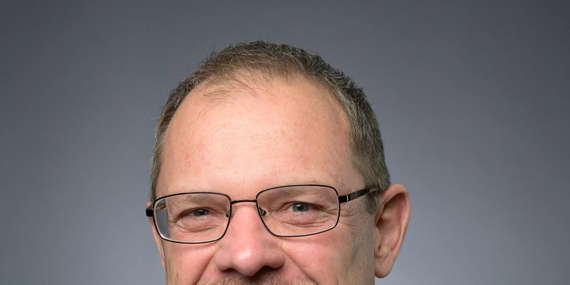 Dwight Scott, president and CEO of the Saint Louis Zoo, in a studio portrait wearing a dark suit, white shirt and blue patterned tie, with glasses and a short gray beard, against a gray background.