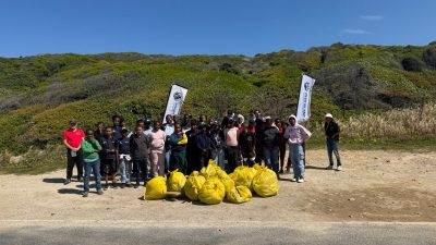 A large group of schoolchildren and adults standing on a sandy roadside below green coastal fynbos behind a pile of about a dozen full yellow refuse bags, with two white banners flying.
