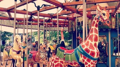 Carved wooden animals on the Birmingham Zoo carousel, including a tall giraffe with a red bridle, a spotted leopard, a zebra, an ostrich and two pale horses, under a lit wooden canopy with species names painted on the boards above.