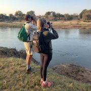 Two people seen from behind, standing on a grassy bank above a wide brown river with day packs on and binoculars raised, looking across at a low far bank of sand, scrub and scattered trees under a pale early morning sky.