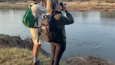 Two people seen from behind, standing on a grassy bank above a wide brown river with day packs on and binoculars raised, looking across at a low far bank of sand, scrub and scattered trees under a pale early morning sky.