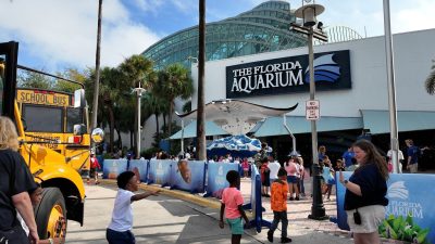 Schoolchildren step off a yellow school bus and walk toward the entrance of The Florida Aquarium in Tampa, past a stingray sculpture beneath the building glass atrium.