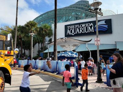 Schoolchildren step off a yellow school bus and walk toward the entrance of The Florida Aquarium in Tampa, past a stingray sculpture beneath the building glass atrium.