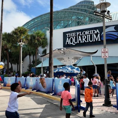 Schoolchildren step off a yellow school bus and walk toward the entrance of The Florida Aquarium in Tampa, past a stingray sculpture beneath the building glass atrium.