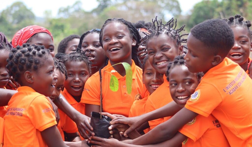 A crowd of girls in bright orange polo shirts stand close together outdoors, laughing, with a dozen hands reaching in to hold a black planting bag containing a young tree seedling with broad green leaves. The shirts carry printed Portuguese text about girls' rights and ending early marriage, and a circular Gorongosa badge on the sleeve. Trees and open sky are behind them.