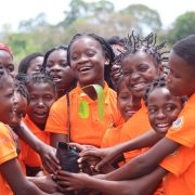 A crowd of girls in bright orange polo shirts stand close together outdoors, laughing, with a dozen hands reaching in to hold a black planting bag containing a young tree seedling with broad green leaves. The shirts carry printed Portuguese text about girls' rights and ending early marriage, and a circular Gorongosa badge on the sleeve. Trees and open sky are behind them.