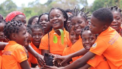 A crowd of girls in bright orange polo shirts stand close together outdoors, laughing, with a dozen hands reaching in to hold a black planting bag containing a young tree seedling with broad green leaves. The shirts carry printed Portuguese text about girls' rights and ending early marriage, and a circular Gorongosa badge on the sleeve. Trees and open sky are behind them.