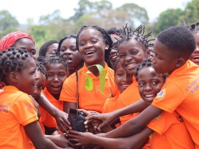 A crowd of girls in bright orange polo shirts stand close together outdoors, laughing, with a dozen hands reaching in to hold a black planting bag containing a young tree seedling with broad green leaves. The shirts carry printed Portuguese text about girls' rights and ending early marriage, and a circular Gorongosa badge on the sleeve. Trees and open sky are behind them.