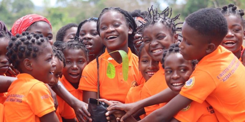 A crowd of girls in bright orange polo shirts stand close together outdoors, laughing, with a dozen hands reaching in to hold a black planting bag containing a young tree seedling with broad green leaves. The shirts carry printed Portuguese text about girls' rights and ending early marriage, and a circular Gorongosa badge on the sleeve. Trees and open sky are behind them.