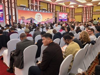 A conference hall filled with delegates seated in white-draped chairs, facing stage screens showing an African savanna graphic and the word WELCOME.