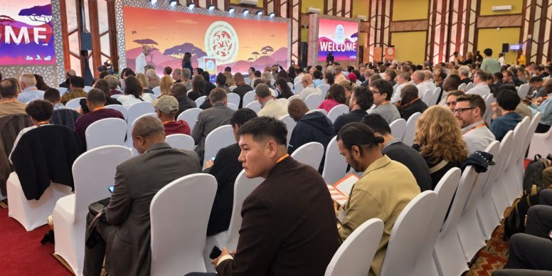 A conference hall filled with delegates seated in white-draped chairs, facing stage screens showing an African savanna graphic and the word WELCOME.