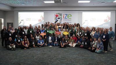 A large group of conference delegates stands and crouches together for a group photograph in front of a screen reading IZE International Zoo Educators Conference Cape Town 2026.