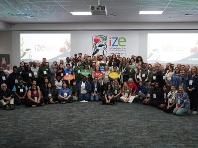 A large group of conference delegates stands and crouches together for a group photograph in front of a screen reading IZE International Zoo Educators Conference Cape Town 2026.
